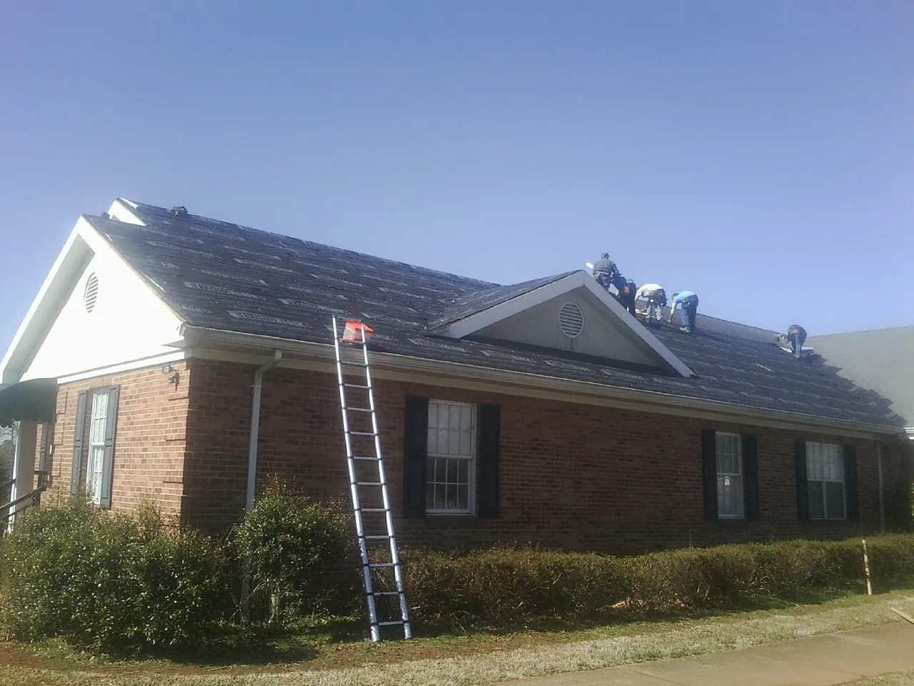 Crew tearing shingles off a brick ranch roof with a ladder set — Metal Roof Repair work by our crew in West Branch