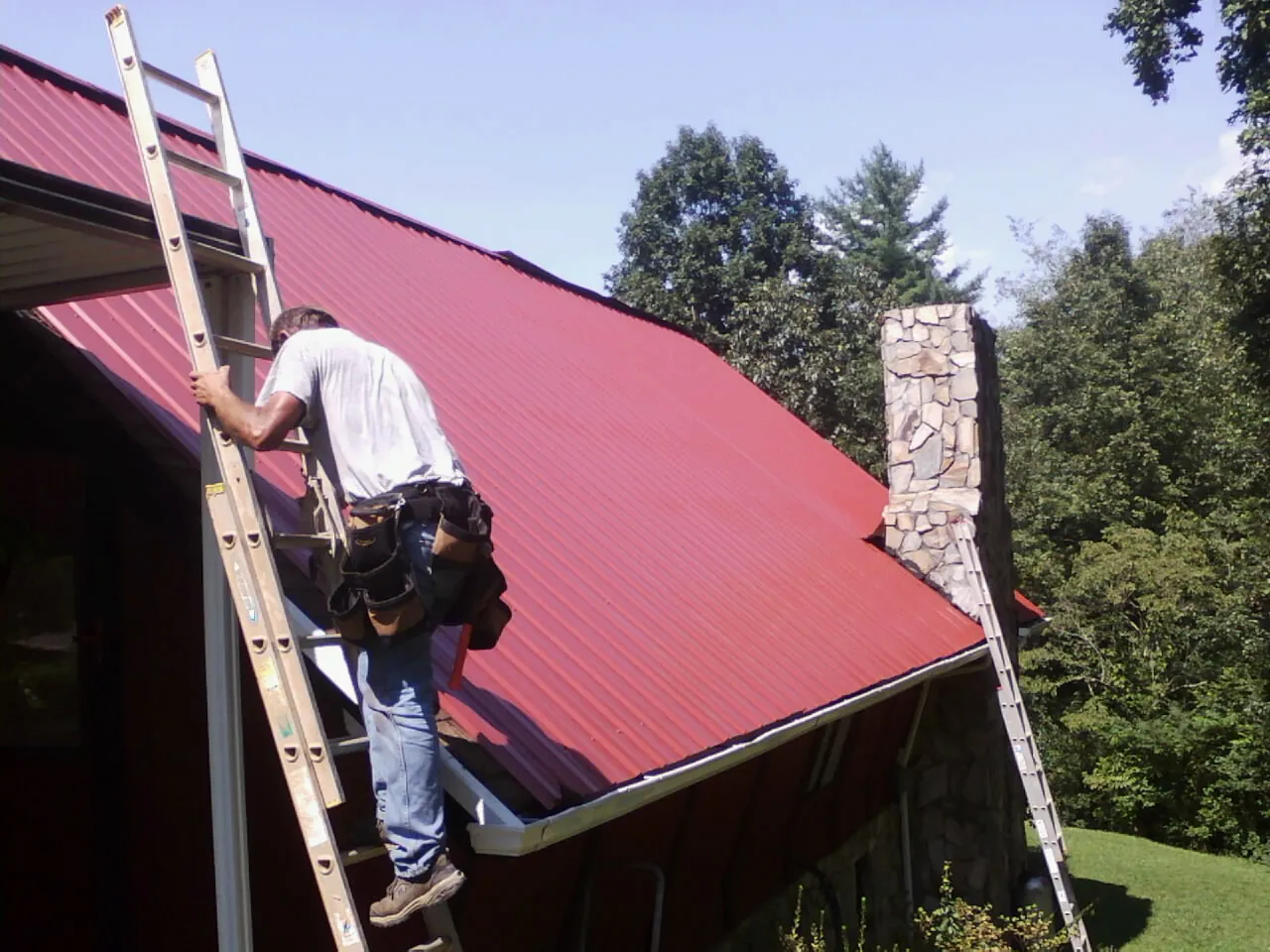 Roofer on a ladder beside a red metal roof and stone chimney — Soffit & Fascia Repair by Roofing Brothers in West Branch